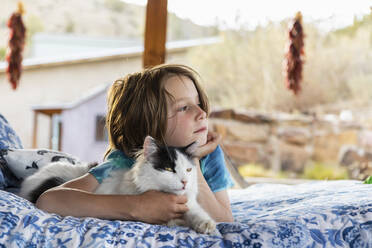 Young boy lying on outdoor bed stroking a pet cat - MINF16280