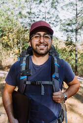 Bearded male backpacker in cap walking among trees and plants in woods in sunny day - ADSF25732