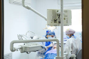 Anonymer Zahnarzt mit einem Kollegen in Uniform, der die Zähne eines Patienten mit medizinischen Instrumenten in einer modernen Klinik operiert - ADSF25738