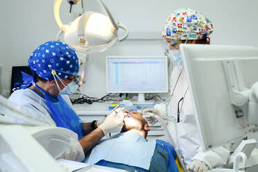 Anonymous dentist with colleague in uniforms operating teeth of patient using medical tools in modern clinic - ADSF25739