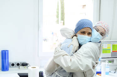 Lächelnde Chirurginnen in sterilen Masken und Uniformen, die sich bei der Arbeit in einer Klinik vor einem Fenster umarmen - ADSF25745
