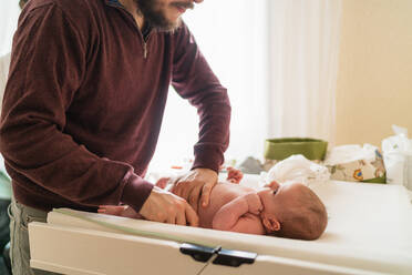 Crop anonymous dad putting diaper on charming little child on infant table in house - ADSF25772