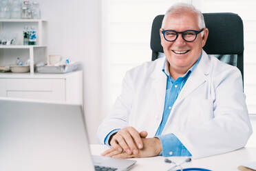 Glücklich lächelnder älterer männlicher Arzt in Uniform und Brille tippt auf einem Netbook am Tisch in einer Klinik - ADSF25777