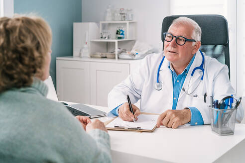 Älterer männlicher Arzt in Uniform und mit Brille füllt ein Formular auf einem Klemmbrett am Tisch gegenüber einer nicht erkennbaren Frau während einer Untersuchung in einer Klinik aus - ADSF25782
