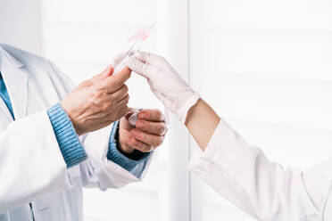 Cropped unrecognizable female coworkers in medical uniforms and sterile masks talking while filling injector with vaccine in hospital - ADSF25785