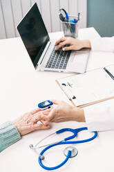 Crop unrecognizable doctor measuring blood oxygen of patient with heart rate monitor at table with stethoscope in clinic while using laptop - ADSF25787