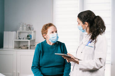 Female medic in uniform with tablet speaking with senior woman in sterile mask on consultation while looking at each other during coronavirus pandemic - ADSF25789