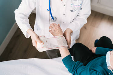 High angle of crop anonymous doctor in uniform wrapping gauze around wrist of patient in hospital - ADSF25794