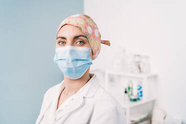 Junge Sanitäterin in weißer Uniform und mit Stoffmütze mit Ornament, die in die Kamera schaut, auf zweifarbigem Hintergrund während einer Coronavirus-Pandemie - ADSF25805