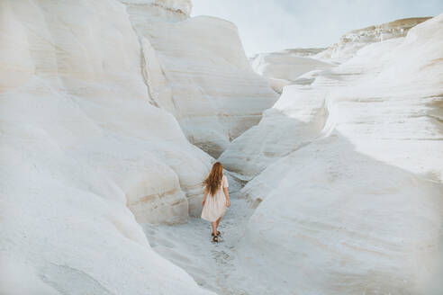 Back view anonymous female in white sundress strolling along narrow passage formed by light curvy rocks on sunny weather in Sarakiniko Greece - ADSF25837