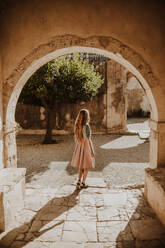 Back view unrecognizable female in stylish summer dress standing near medieval building arch on sunny summer day in Heraklion Milos - ADSF25843