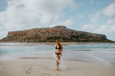 Full length slim female in black swimsuit standing on sandy Balos beach against rocky cliff and looking away on clear sunny weather - ADSF25846