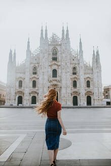 Rückenansicht einer anonymen weiblichen Touristin mit fliegenden Haaren auf dem Bürgersteig vor einer alten gemauerten Kathedrale mit Dekor in Mailand Italien - ADSF25848