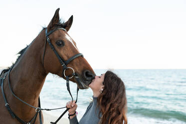 Side view of young female kissing muzzle of chestnut stallion in bridle against wavy ocean - ADSF25857