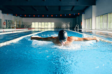 Starker männlicher Schwimmer mit Badekappe beim Schmetterlingsschlag während des Trainings im Schwimmbad mit blauem Wasser - ADSF25871