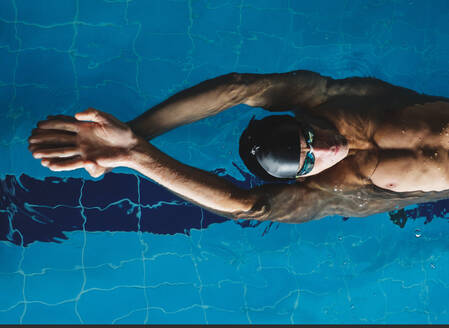 Top view of male athlete in bathing cap with raised arms swimming on back in pool during training - ADSF25873