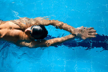 Draufsicht auf einen männlichen Athleten mit Badekappe und erhobenen Armen, der während des Trainings auf dem Rücken schwimmt - ADSF25874