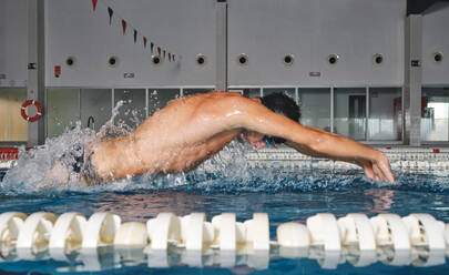 Seitenansicht eines starken männlichen Schwimmers mit Badekappe, der während des Trainings im Schwimmbad mit blauem Wasser Schmetterlingsschwimmen betreibt - ADSF25875