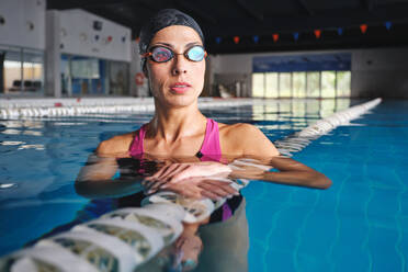 Erwachsene Sportlerin in Schwimmbrille und Badekleidung, die sich auf eine Bahn im Schwimmbad mit transparentem Wasser stützt und wegschaut - ADSF25877