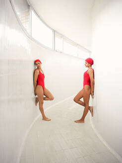 Side view of young female athletes in same swimwear with raised legs standing on tiled floor in passage - ADSF25885