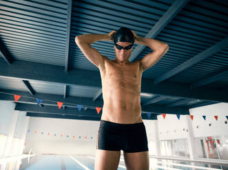 Sportler in Badehose und Kappe mit den Händen hinter dem Kopf bei der Vorbereitung auf das Training im Schwimmbad mit Bahnen im Gebäude - ADSF25893