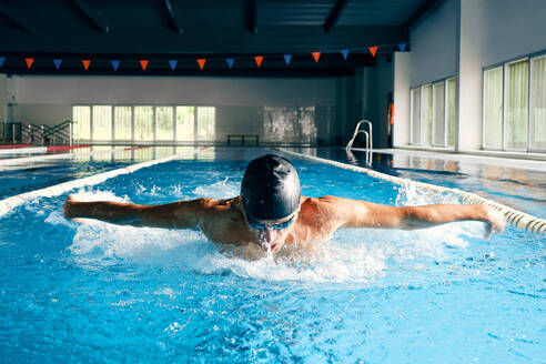 Strong male swimmer in bathing cap performing butterfly stroke during workout in swimming pool with blue water - ADSF25894