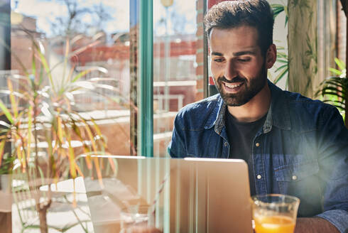 Blick durch Glas auf einen erwachsenen, bärtigen, ethnischen, männlichen Angestellten, der an einem Tisch an einem Netbook arbeitet - ADSF25905