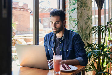 Side view of ethnic male employee working on netbook at table with tasty cake piece and drinks - ADSF25907