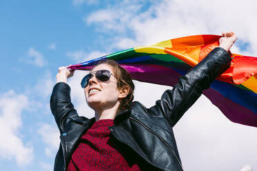 Von unten entzückte lesbische Frau, die auf der Straße mit einer im Wind flatternden LGBT-Regenbogenfahne steht und gegen den bewölkten Himmel wegschaut - ADSF25918