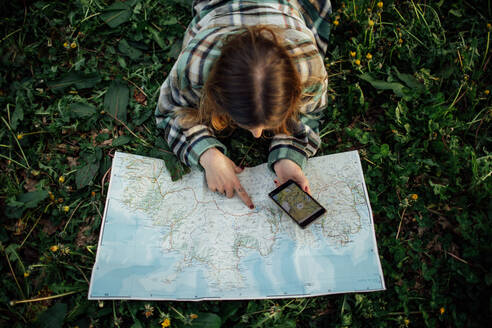 From above back view of unrecognizable female tourist with cellphone watching route map while lying on grass in countryside - ADSF25926