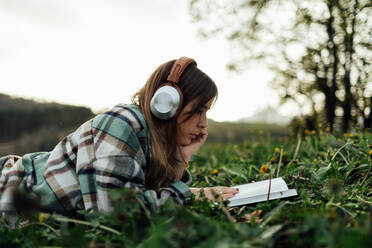 Seitenansicht einer jungen aufmerksamen Frau mit modernem Headset, die im Sommer auf einer Wiese liegend ein Lehrbuch liest - ADSF25927