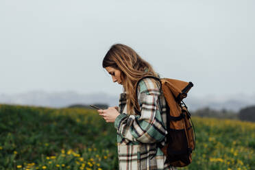 Seitenansicht einer Wanderin, die eine GPS-Anwendung auf einem Mobiltelefon benutzt, während sie auf einer Wiese gegen die Berge läuft - ADSF25929