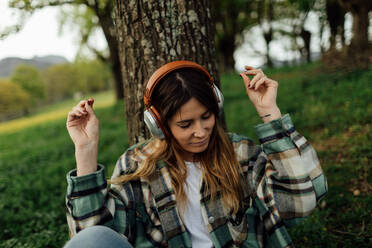 Young content female with tattoo in checkered shirt listening to music from headphones on lawn in summer - ADSF25931