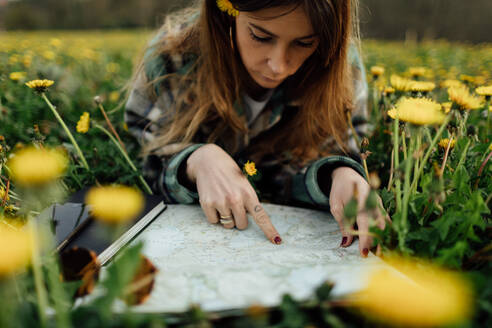 Concentrated female traveler with paper map and blossoming flowers looking away while lying on meadow against mountain in countryside - ADSF25932