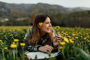 Content female traveler with paper map and blossoming flowers looking away while lying on meadow against mountain in countryside - ADSF25933