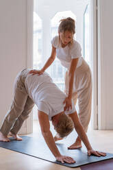 Mann übt Yoga in Downward Facing Dog Pose mit Hilfe einer Lehrerin während einer Sitzung zu Hause - ADSF25938