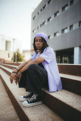 Ganzkörperaufnahme einer ernsten afroamerikanischen Frau in Freizeitkleidung und mit Kopftuch, die in die Kamera schaut, während sie auf Steinstufen in der Nähe eines Gebäudes in der Stadt sitzt - ADSF25954