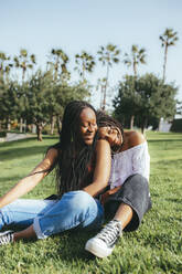 Ganzkörperaufnahme von ruhigen afroamerikanischen Freundinnen, die sich aneinander kuscheln und in die Kamera schauen, während sie auf einer Wiese im Park sitzen - ADSF25955