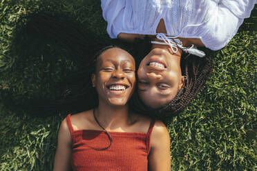 Draufsicht auf lächelnde afroamerikanische Freundinnen, die in die Kamera schauen, während sie an einem sonnigen Sommertag auf einer Wiese liegen - ADSF25957