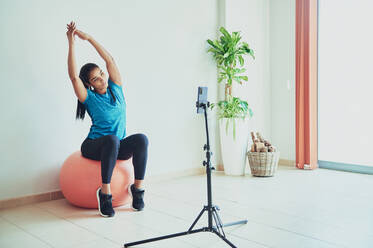 Young ethnic vlogger working out with raised arms on exercise ball while recording video on cellphone in room - ADSF25967