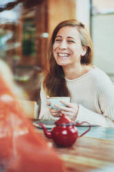 Happy female with cup of tea speaking with crop anonymous friend at table in cafeteria - ADSF25973