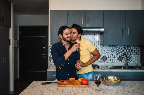 Ethnischer Mann küsst und umarmt homosexuelle Geliebte mit einem Glas Rotwein am Tisch mit Brot und Tomaten zu Hause - ADSF25986