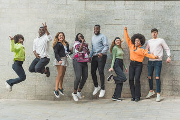 Fröhliche junge multiethnische Partner, die eine Siegesgeste zeigen, während sie über einen Gehweg springen und in die Kamera in einer Stadt auf grauem Hintergrund schauen - ADSF25993