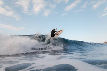 Anonymer männlicher Surfer auf einem Surfbrett bei der Ausübung eines Extremsports auf einer Meereswelle unter bewölktem Himmel - ADSF26008