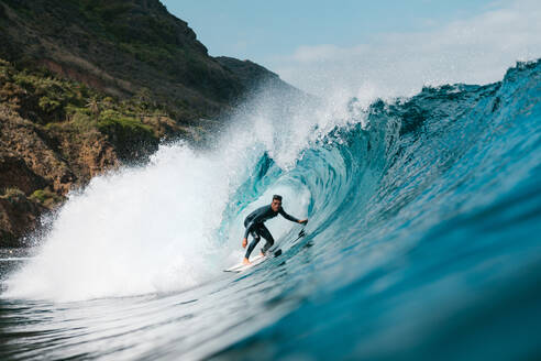 Sportsman in wetsuit on surfboard practicing extreme sport on wavy ocean with splattering aqua against mountain - ADSF26010