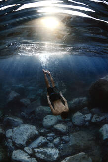 Back view of unrecognizable male athlete swimming under blue ocean water with ripples and boulders on bottom - ADSF26013