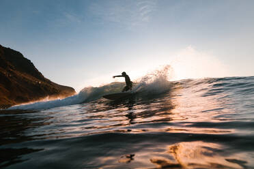 Anonyme männliche Surfer-Silhouette auf einem Surfbrett beim Extremsport auf einer Meereswelle unter bewölktem Himmel bei Sonnenuntergang - ADSF26014