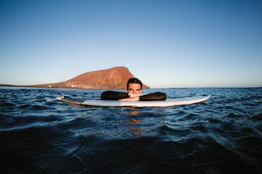 Happy male resting surfer on surfboard in wavy sea looking at camera against mount in daytime - ADSF26015