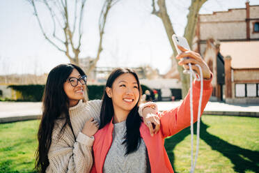 Positive junge ethnische weibliche Freunde mit langen dunklen Haaren in lässiger Kleidung lächelnd und zeigt V-Zeichen bei der Aufnahme von Selfie auf Smartphone auf sonnigen Tag im Park - ADSF26021