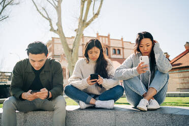 Junge asiatische Freunde in stilvollen Outfits benutzen Smartphones, während sie an einem sonnigen Tag auf einer Steinbrüstung im Stadtpark sitzen - ADSF26025